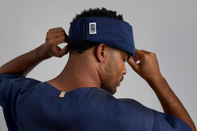 Man wearing a dark blue silk sleep mask over his forehead with a LUNYA logo on a gray background