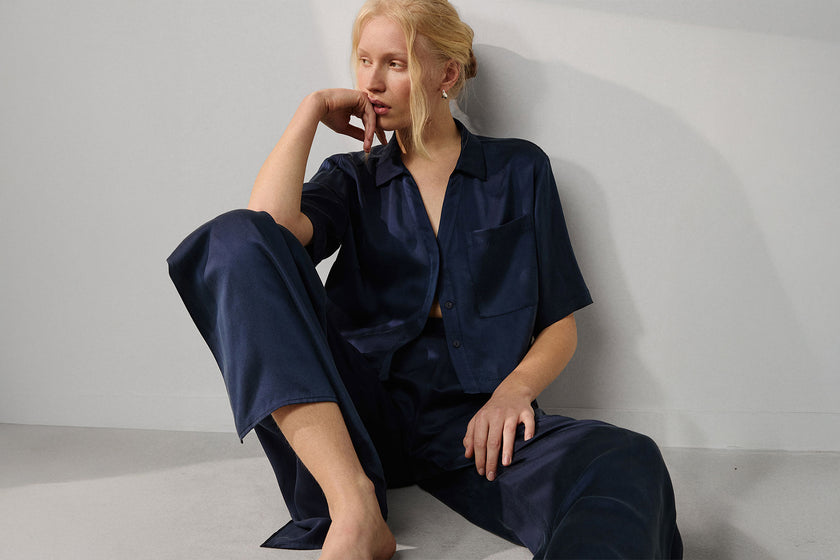 Woman wearing a deep blue silk pajama set sitting against a light gray background