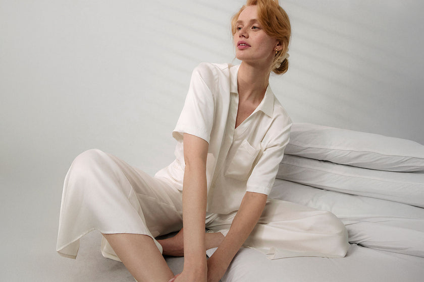 Woman sitting on a bed wearing a white silk pajama set against a plain background