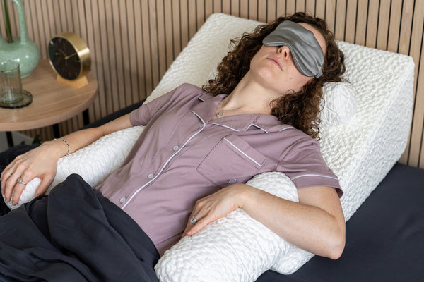 A woman lying in dark bedding with a grey eye mask on and laying against a white wedge pillow with three easy breather bolster pillows under her neck and arms for additional support