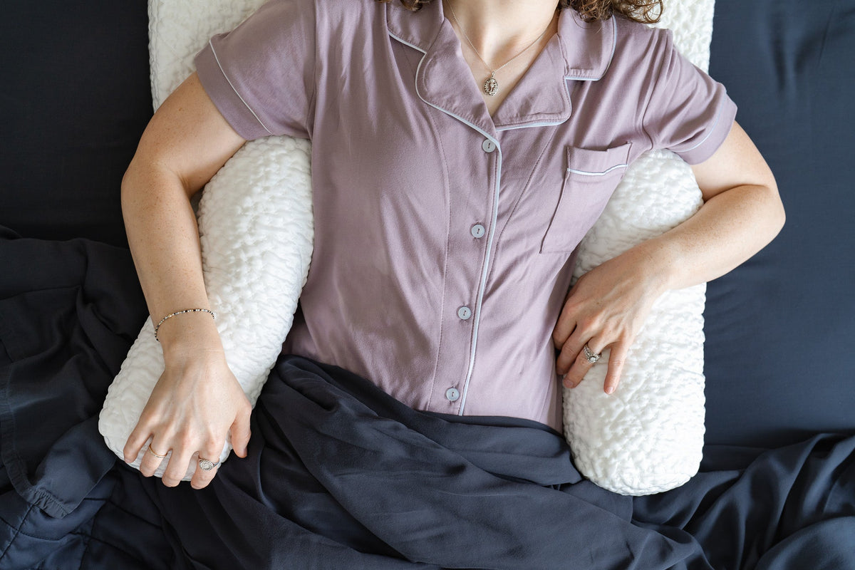Person lying on a bed with two white bolster pillows under her arms and covered in dark bedding