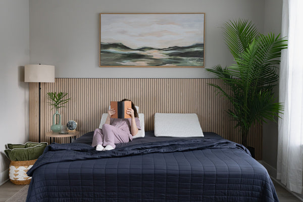 Person reading a book in bed while using an easy breather wedge pillow for support, with a scenic painting on the wall