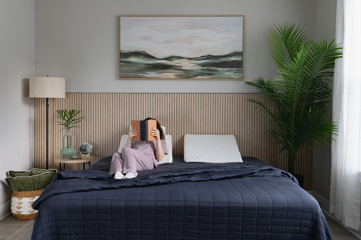 Person reading a book in bed while using an easy breather wedge pillow for support, with a scenic painting on the wall