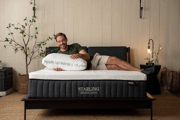 a man joyfully lying on his side while on a starling mattress and holding a plush easy breather pillow