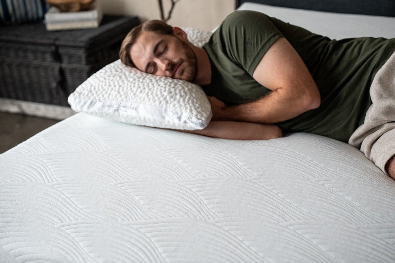 A man lies on his side on a white quilted mattress, resting his head on a white easy breather pillow in a bedroom setting.