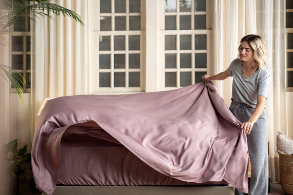 Hotel-inspired home bedroom with woman draping breathable bedding over a bed, soft bedside light, and curtains in the background