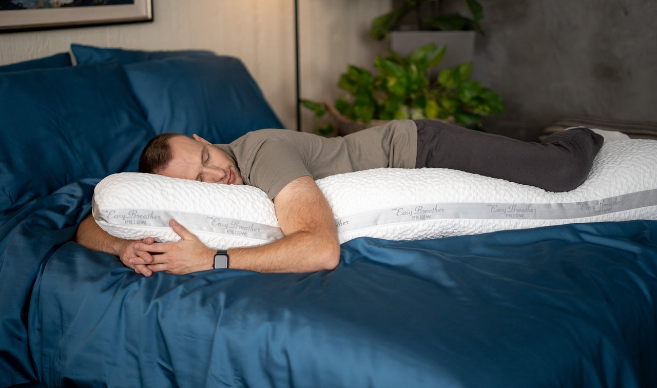 A man laying on a long body pillow arranged for side sleeper support on top of rich blue bamboo bedding
