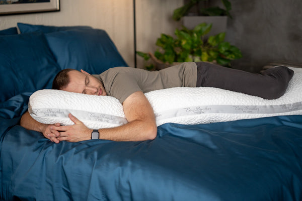 A man lies on his side on a bed with blue bedding, using a white contoured lumbar pillow to support his lower back. The room has a dark gray wall and a green plant in the background.