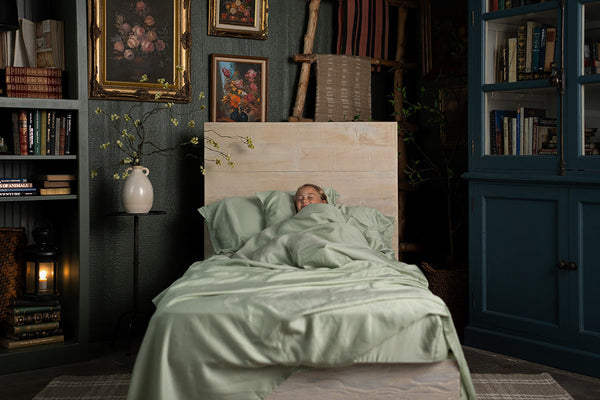 Person lying in bed with green bedding in a dimly lit room with bookshelves and artwork.