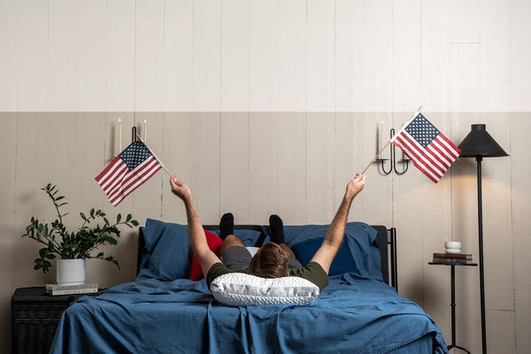 nest bedding bedroom scene, man laying on indigo blue crinkle percale organic cotton sheets and an easy breather jr pillow waving two small american flags in the air