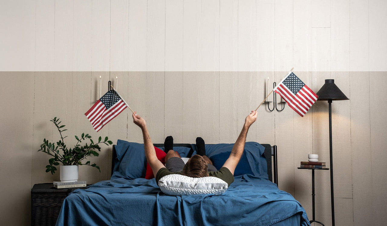 nest bedding bedroom scene, man laying on indigo blue crinkle percale organic cotton sheets and an easy breather jr pillow waving two small american flags in the air