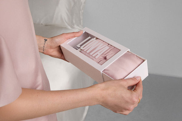 A person wearing pink pajamas holding a LUNYA branded box with a pink silk sleep mask inside