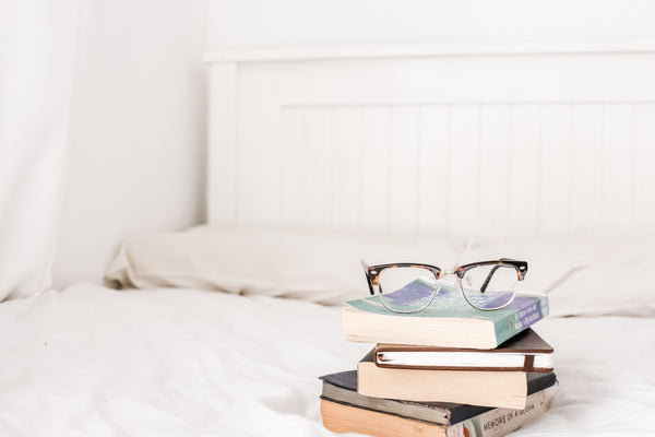 stack of books and eyeglasses on bed with perfect pillows
