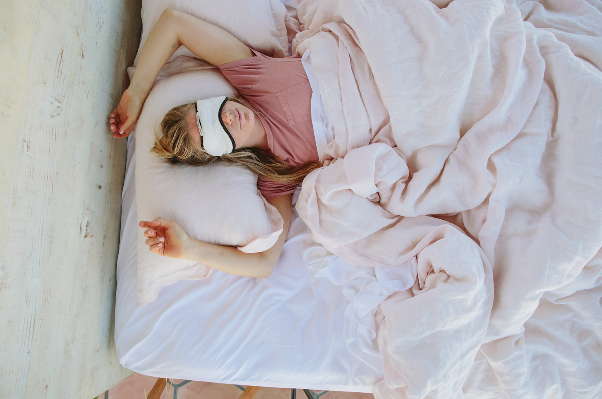 Person sleeping in bed with a white sleep mask on, wearing pink pajamas.