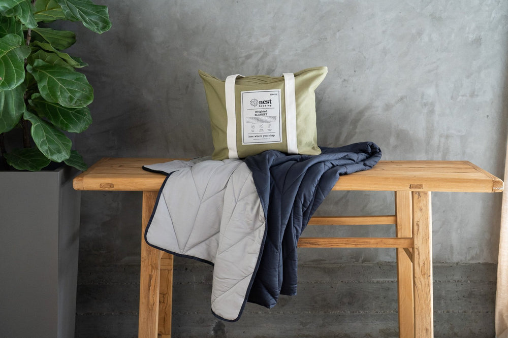 The Luxury Weighted Blanket by Nest Bedding draped on a wooden table with the green cotton tote bag packaging atop the blanket.