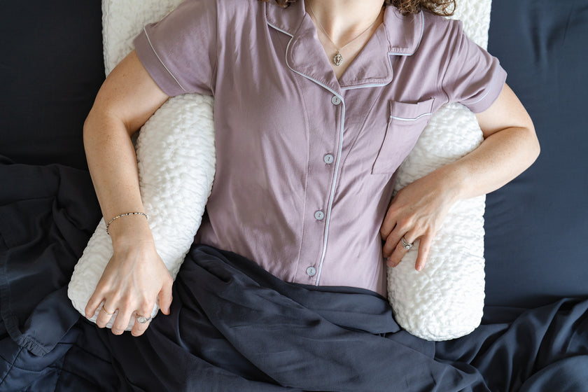 Person lying on a bed with two white bolster pillows under her arms and covered in dark bedding