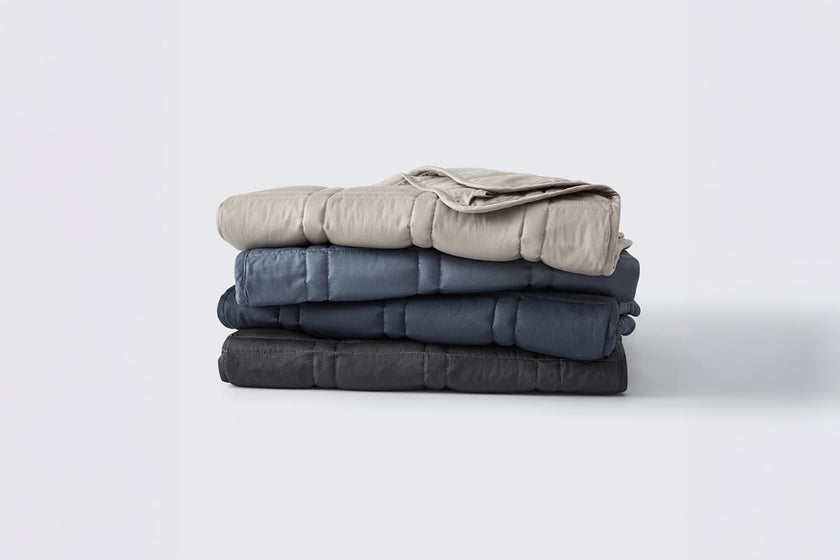 Four folded weighted lap blankets in the colors silver sage, dusk blue, luna blue, and midnight grey, on a white background