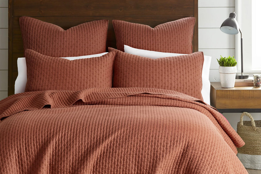 Adobe textured cross stitch quilt set on a bed with pillows and a lamp in the background.