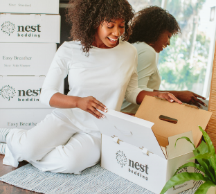 A woman wearing all white looks surprised as she opens a Nest Bedding Easy Breather Pillow box.