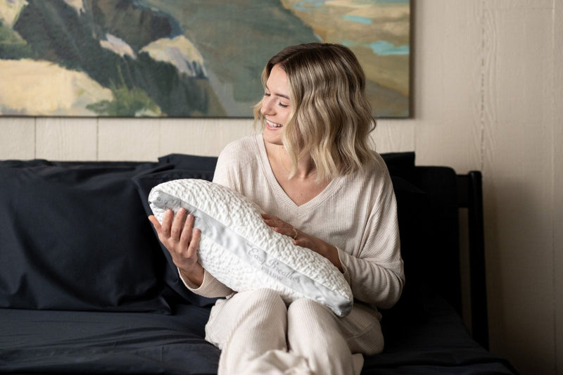 A woman holding a white Easy Breather Jr. pillow in a dark cozy bedroom with a painting on the wall.
