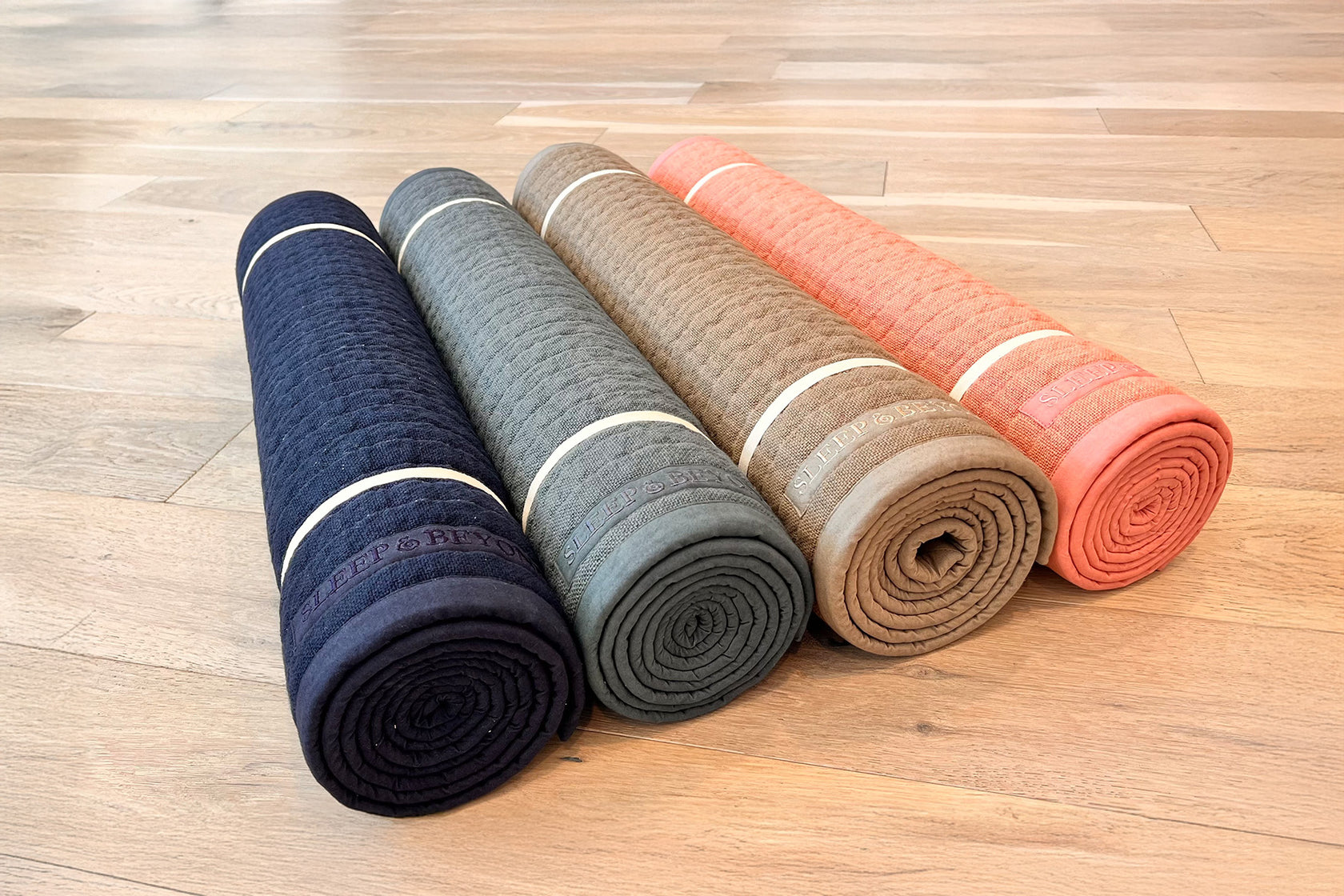 Four rolled-up yoga mats in navy, grey, light brown, and pink on a wooden floor.