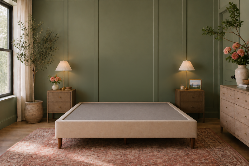 A minimalist upholstered bed frame in a warm beige tone with dark wooden legs, styled in a modern bedroom with sage green paneled walls, matching nightstands, lit table lamps, and a patterned rug.